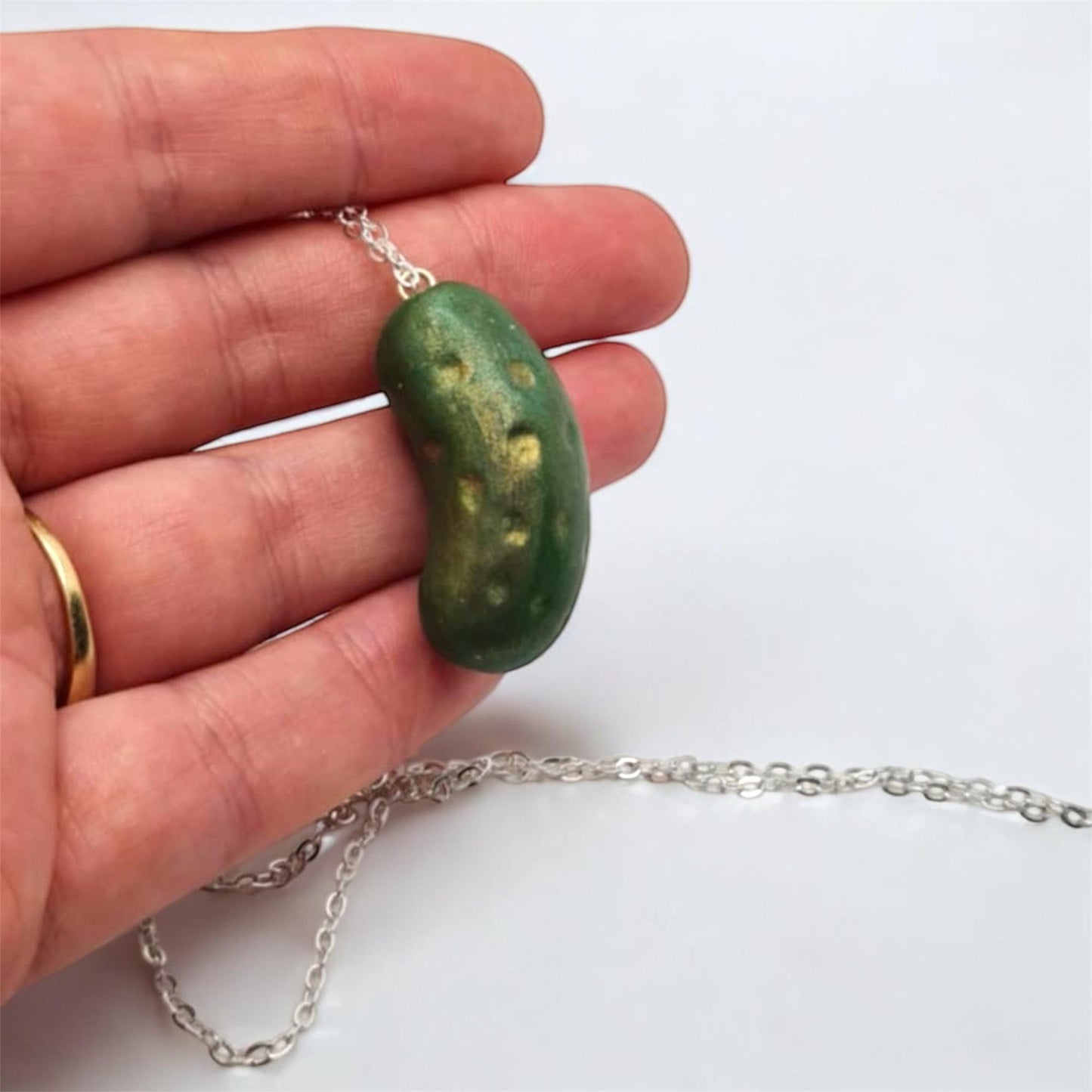 Green pickle pendant on a silver chain held by a hand against a white background