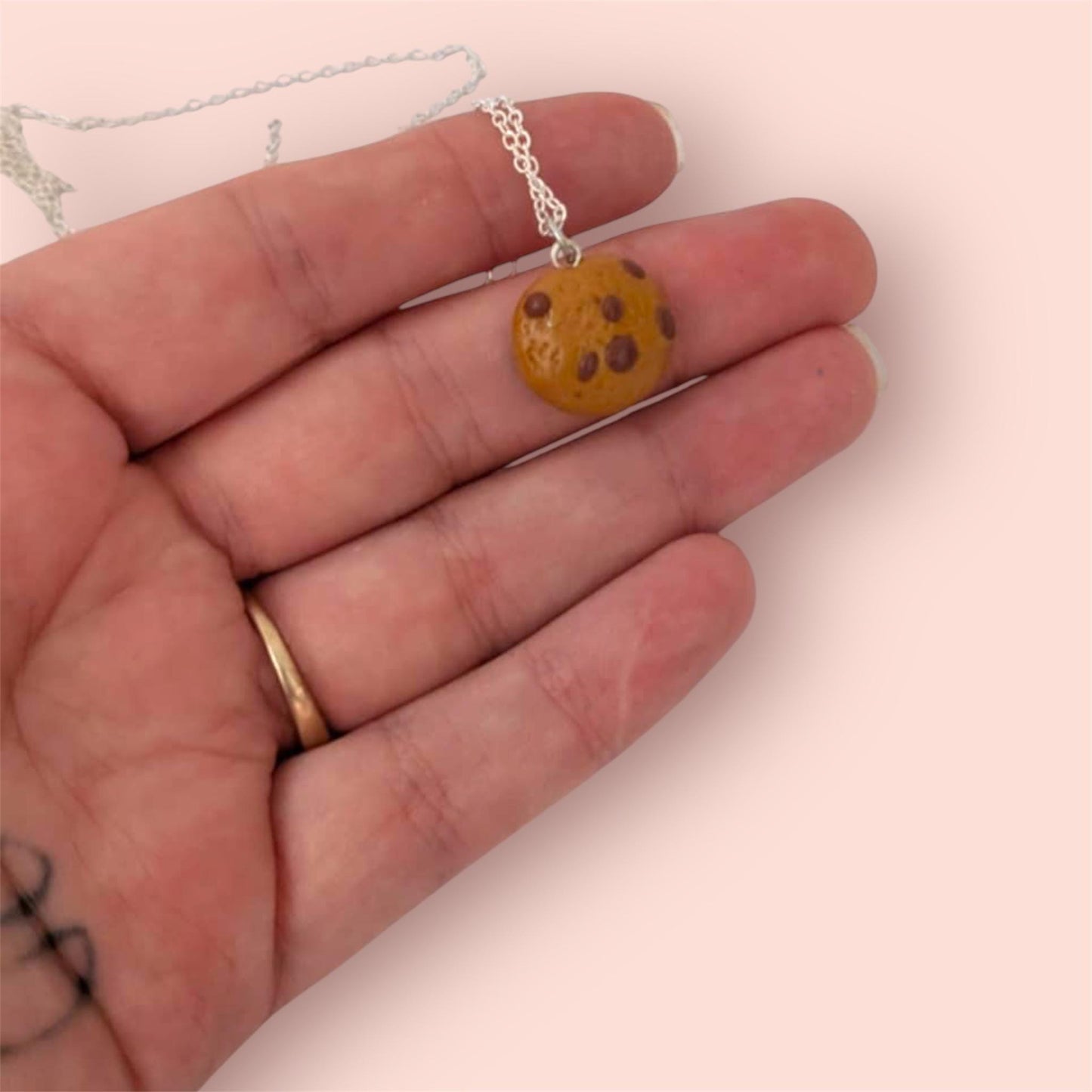 Hand holding a necklace with a cookie-shaped pendant on a light pink background