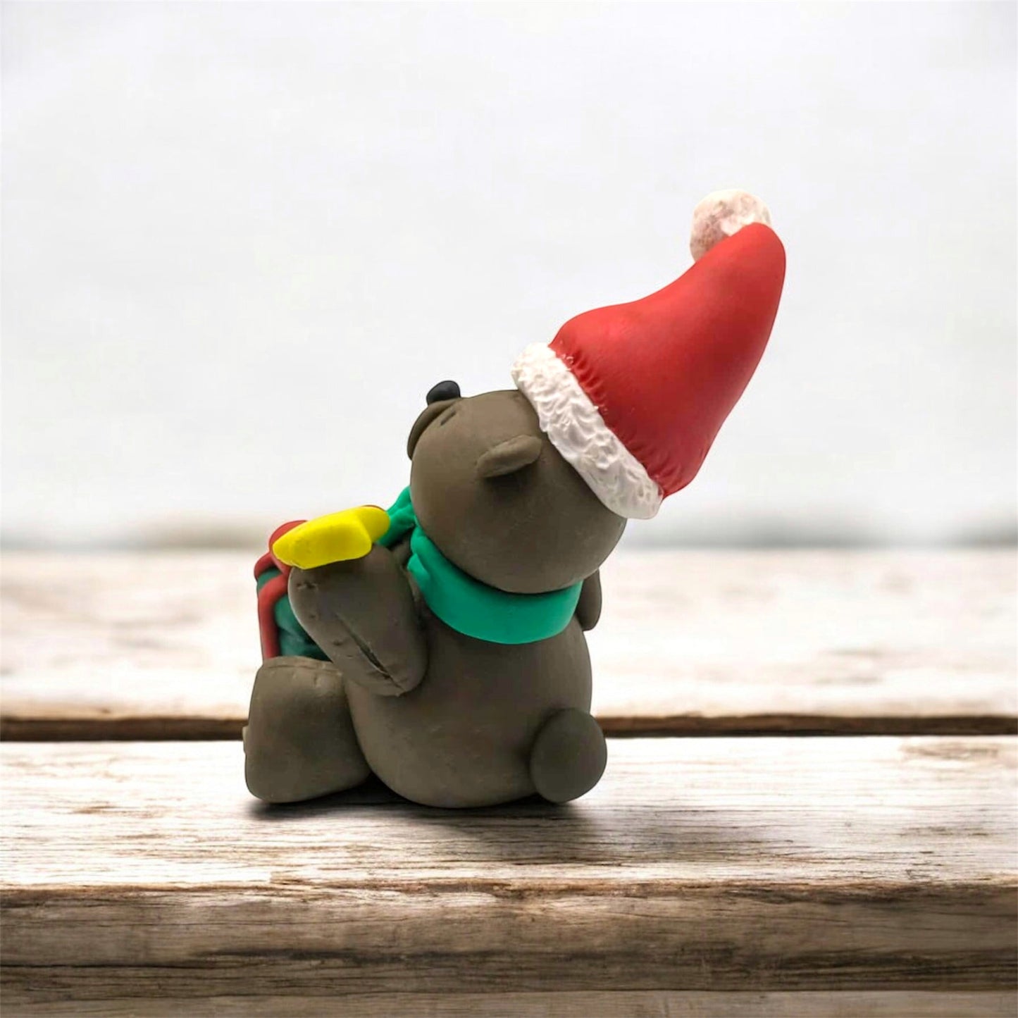 Side and back view of a Small clay figure of a bear wearing a Santa hat on a wooden surface
