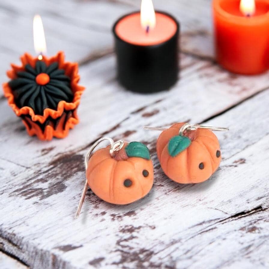 Pumpkin-shaped earrings on a wooden surface with lit candles in the background.