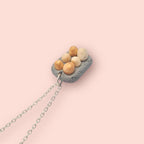 Miniature eggs in a carton on a silver chain against a light pink background 