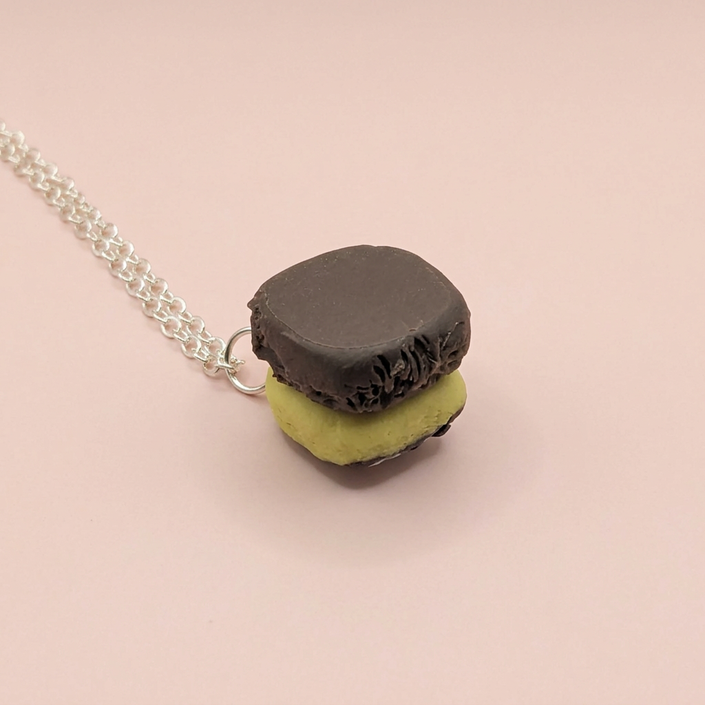 A miniature chocolate traybake necklace chocolate pendant necklace on a delicate silver chain, set against a solid pale pink background.