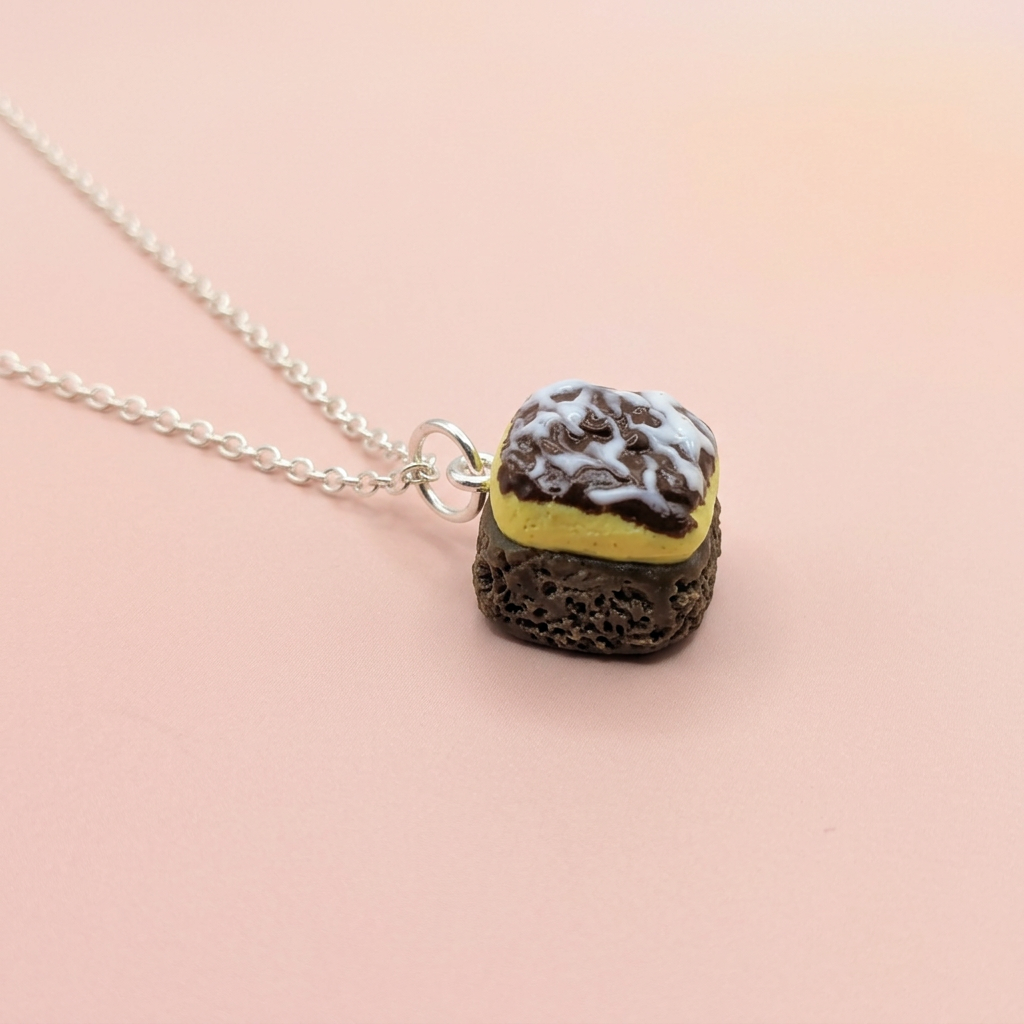 A silver chain necklace with a miniature pendant shaped like a sandwich cookie, featuring a dark brown textured base, a yellow middle layer, and a white top layer with dark speckles, set against a soft pink background. Chocolate traybake necklace 