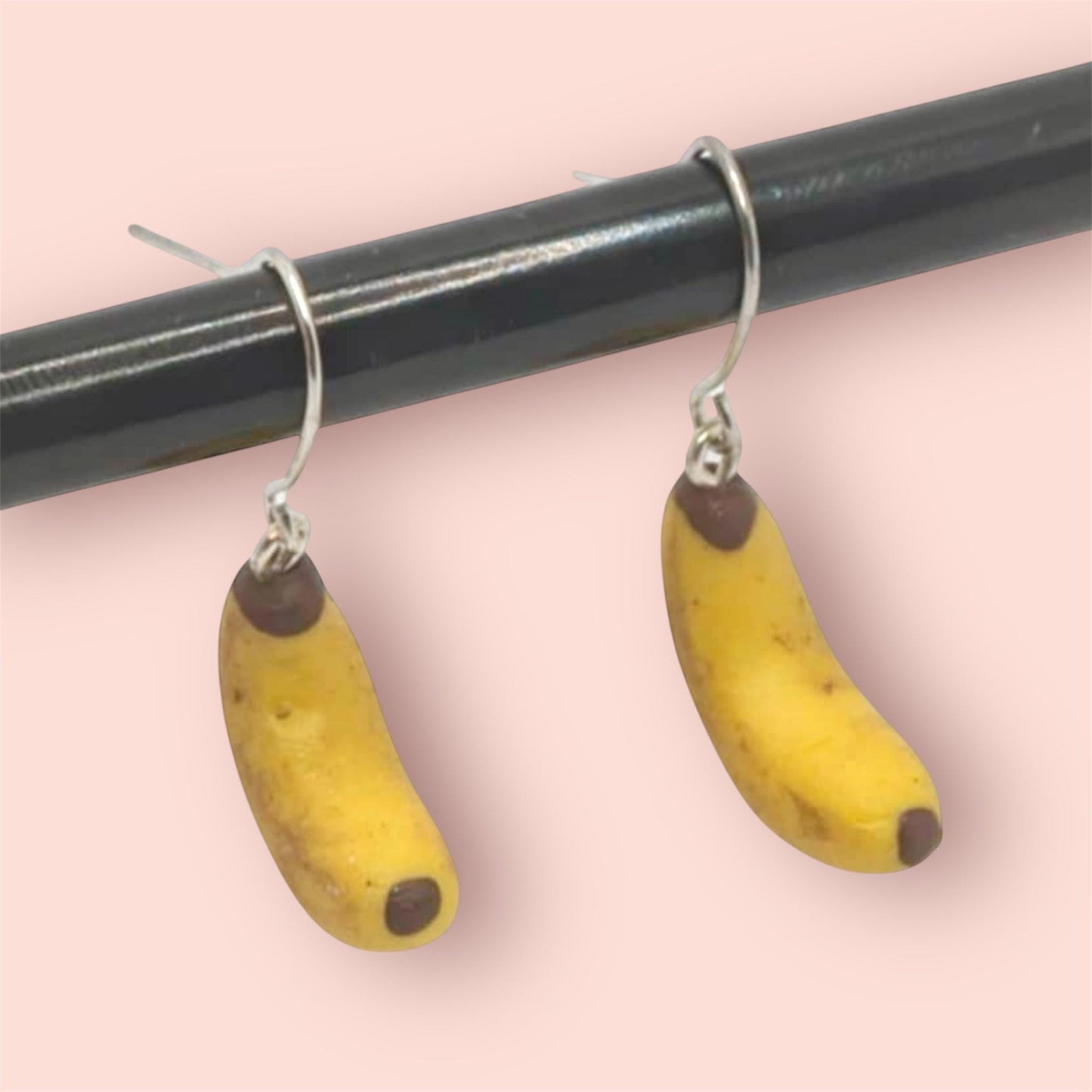 Banana-shaped earrings hanging from a black rod against a pink background