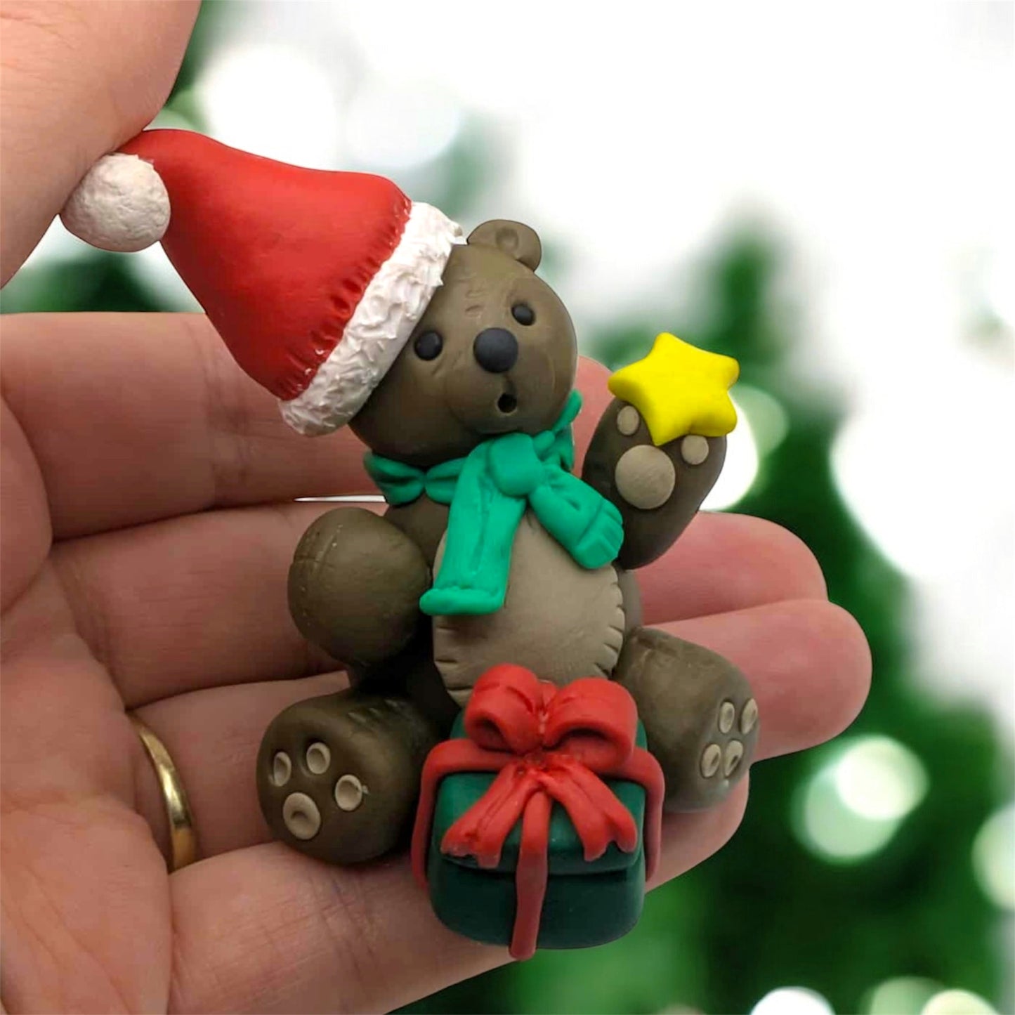Hand holding a small teddy bear figurine with a Santa hat, green scarf, and gift box against a blurred Christmas tree background.