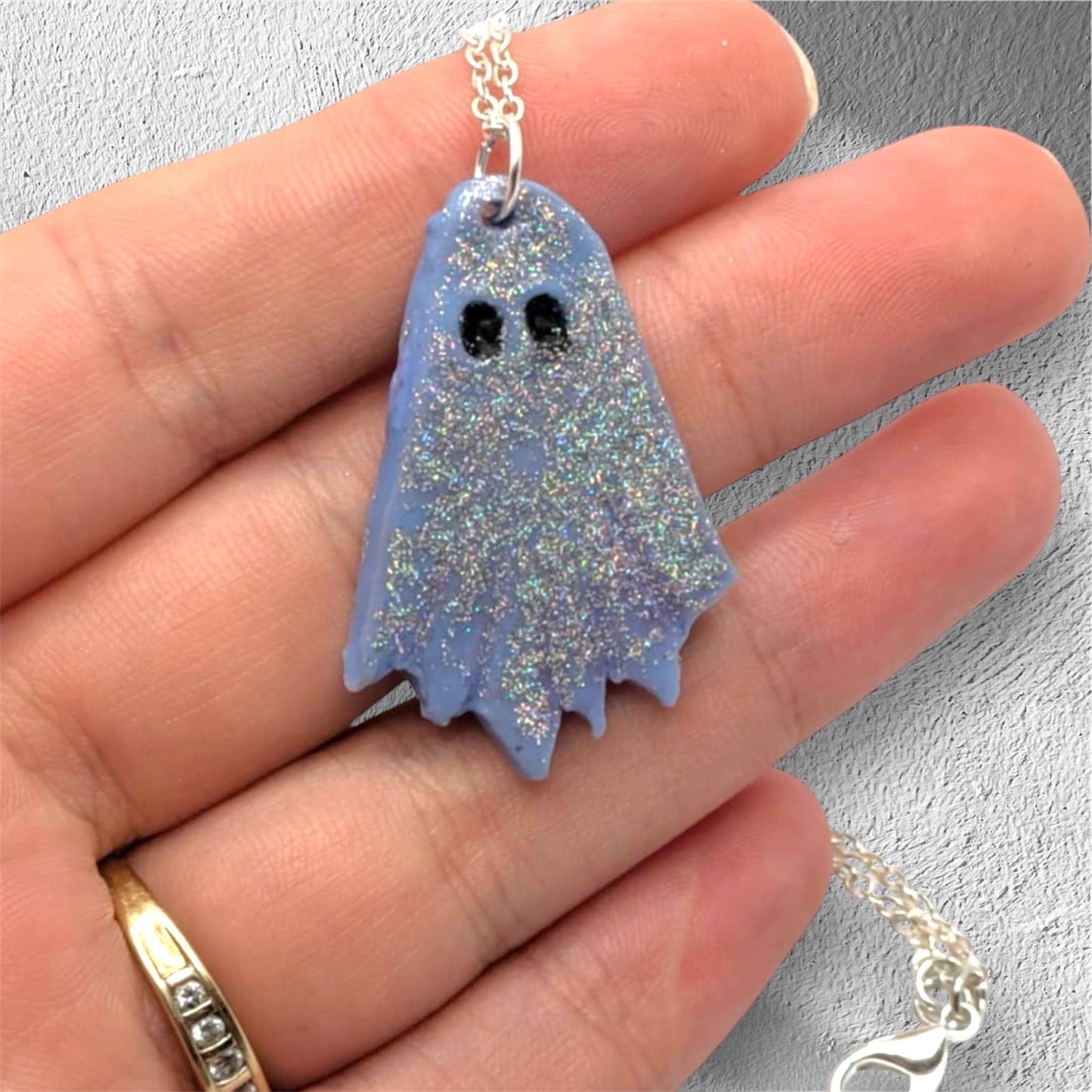 Ghost-shaped pendant on a chain held in a hand against a gray background