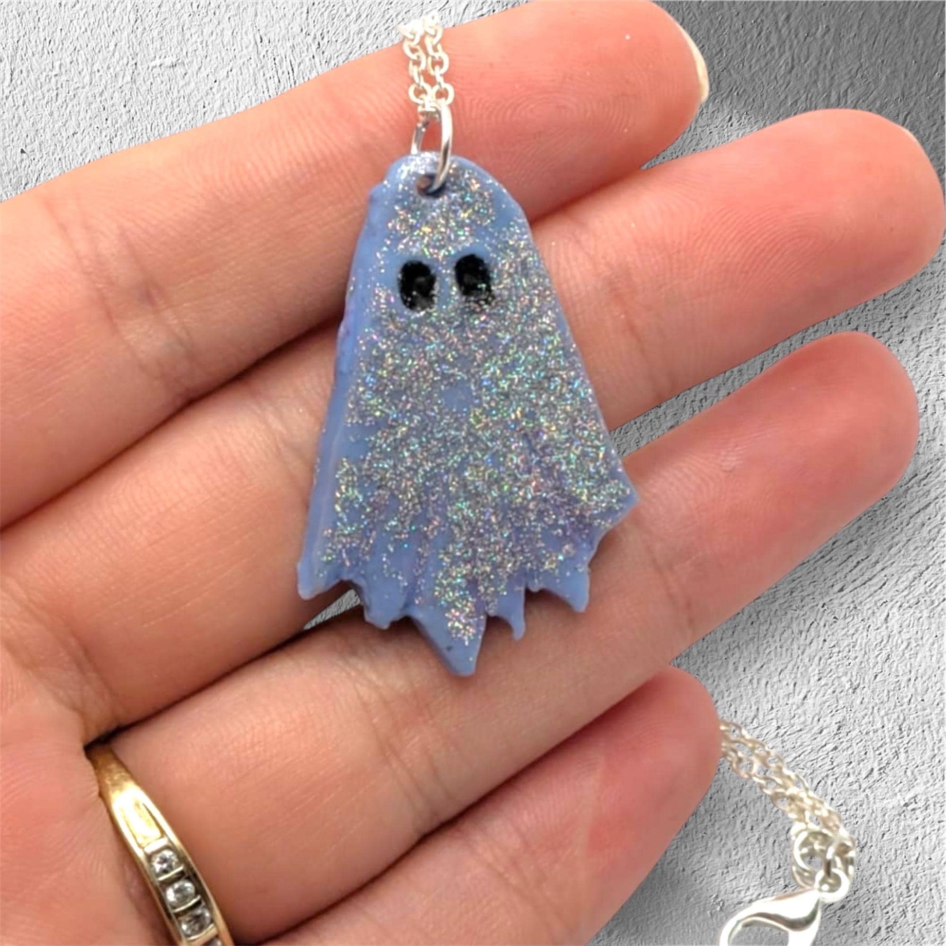 Ghost-shaped pendant on a chain held in a hand against a gray background