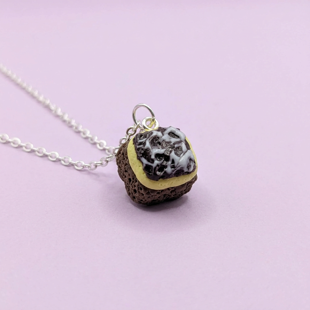 A novelty Chocolate traybake necklace featuring a small pendant shaped like a lava rock cookie with white frosting, resting on a light pink background.