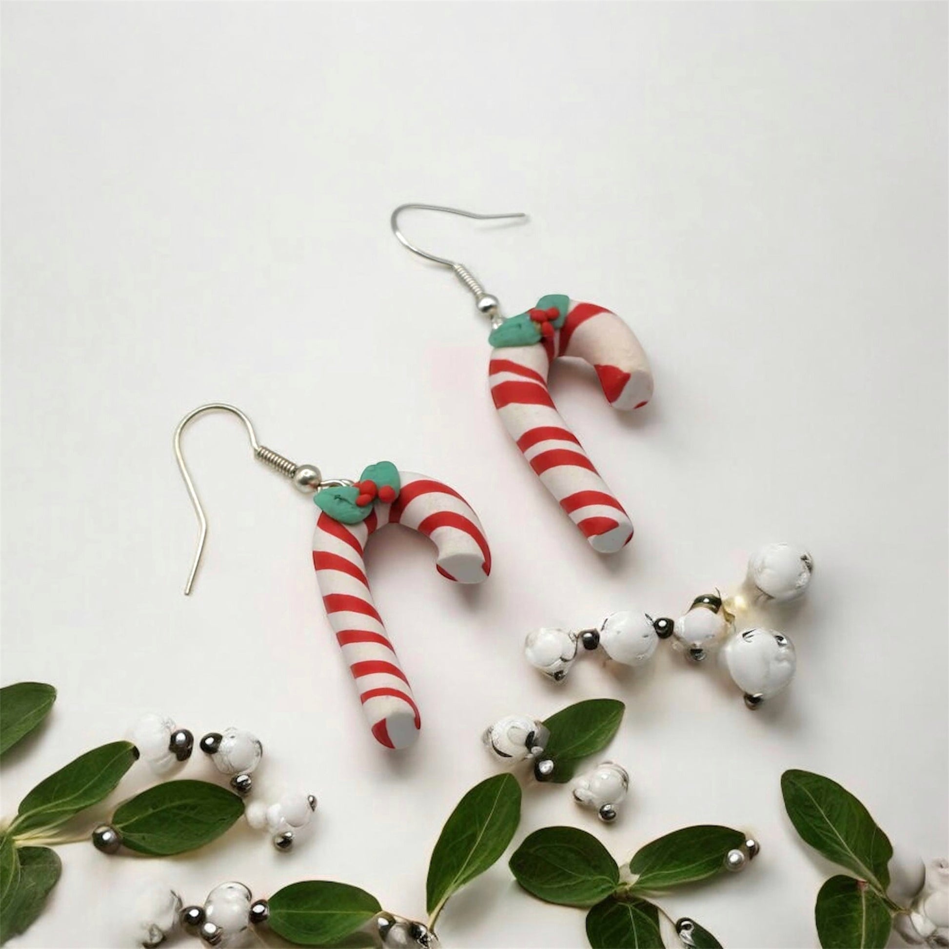 Candy cane-shaped earrings on a white background with green leaves and white berries.