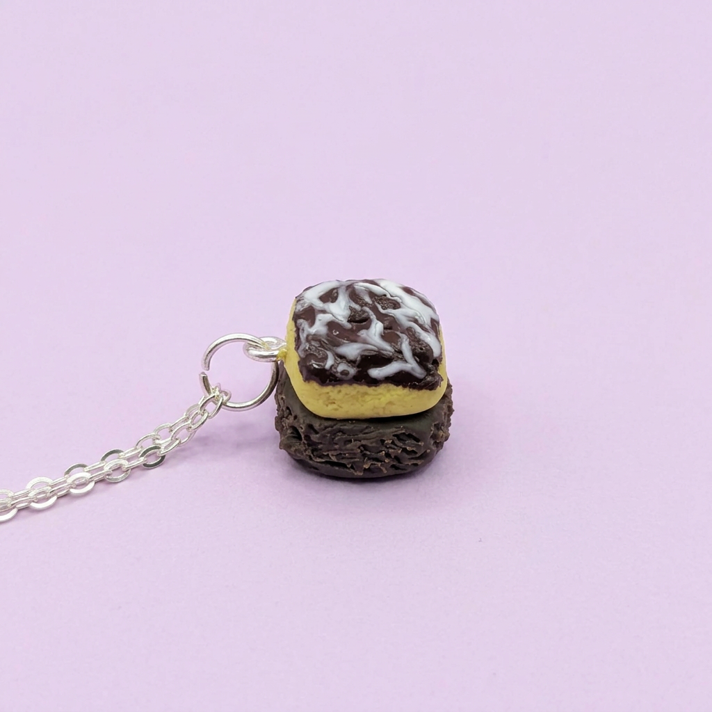 A miniature charm necklace shaped like a chocolate sandwich cookie with yellow frosting and white drizzle, attached to a silver chain against a solid light purple background. Chocolate traybake necklace 