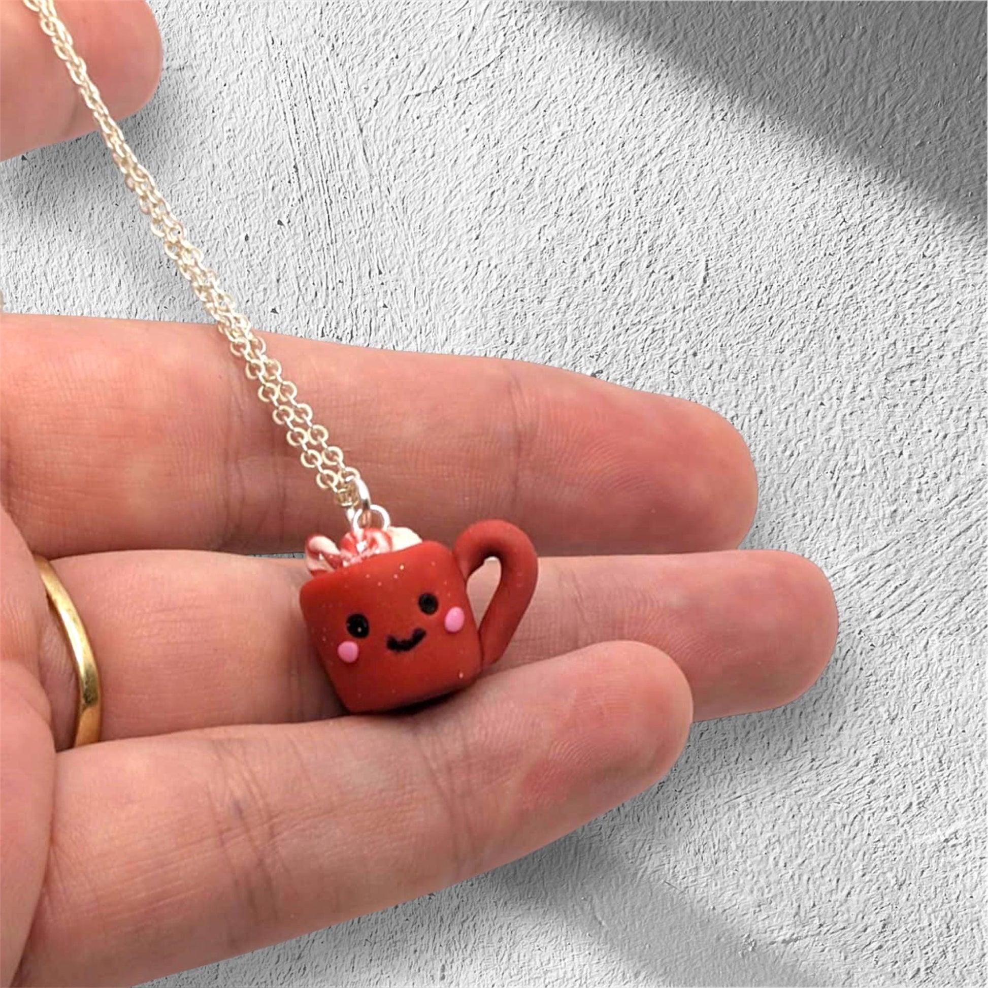 Small red mug pendant with a smiling face on a chain held between fingers against a light gray background.