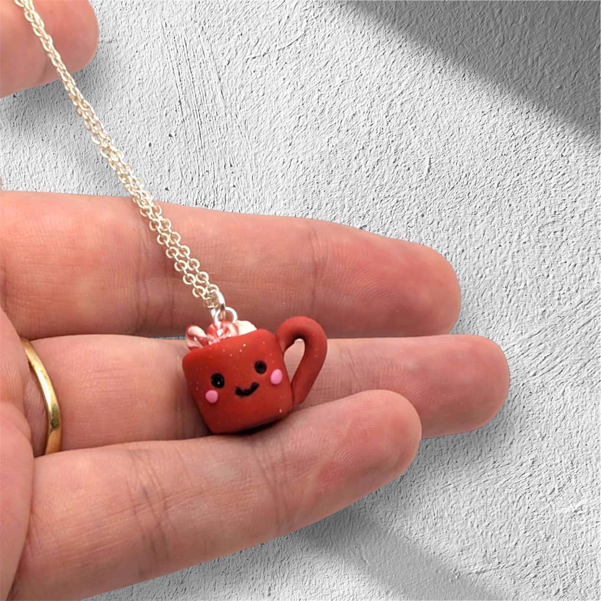 Small red mug pendant with a smiling face on a chain held between fingers against a light gray background.