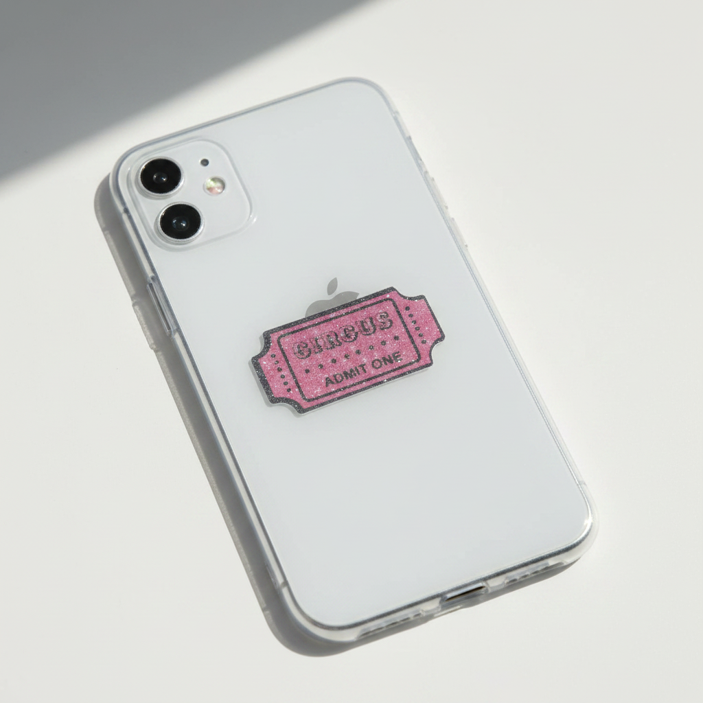 A clear phone case on a white smartphone, featuring a pink vintage ticket charm with 'CIRCUS ADMIT ONE' text, photographed on a clean white background.