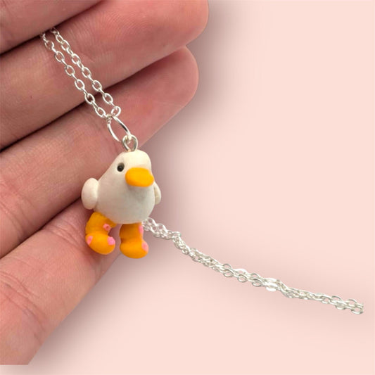 Small duck-shaped pendant on a silver chain held by a hand against a light pink background