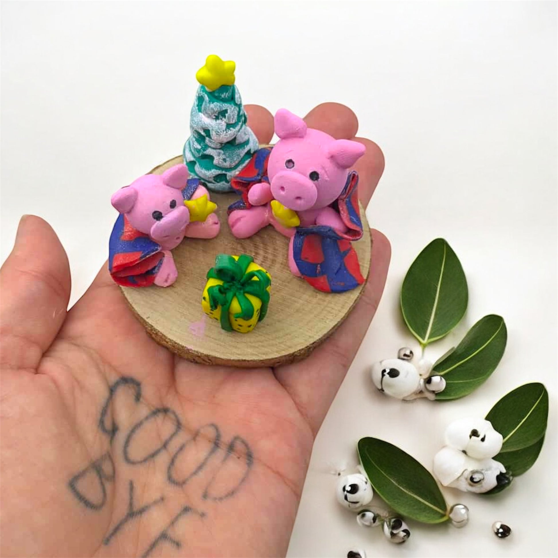 Hand holding small clay figurines on a wooden base with additional figurines and leaves on a white background.