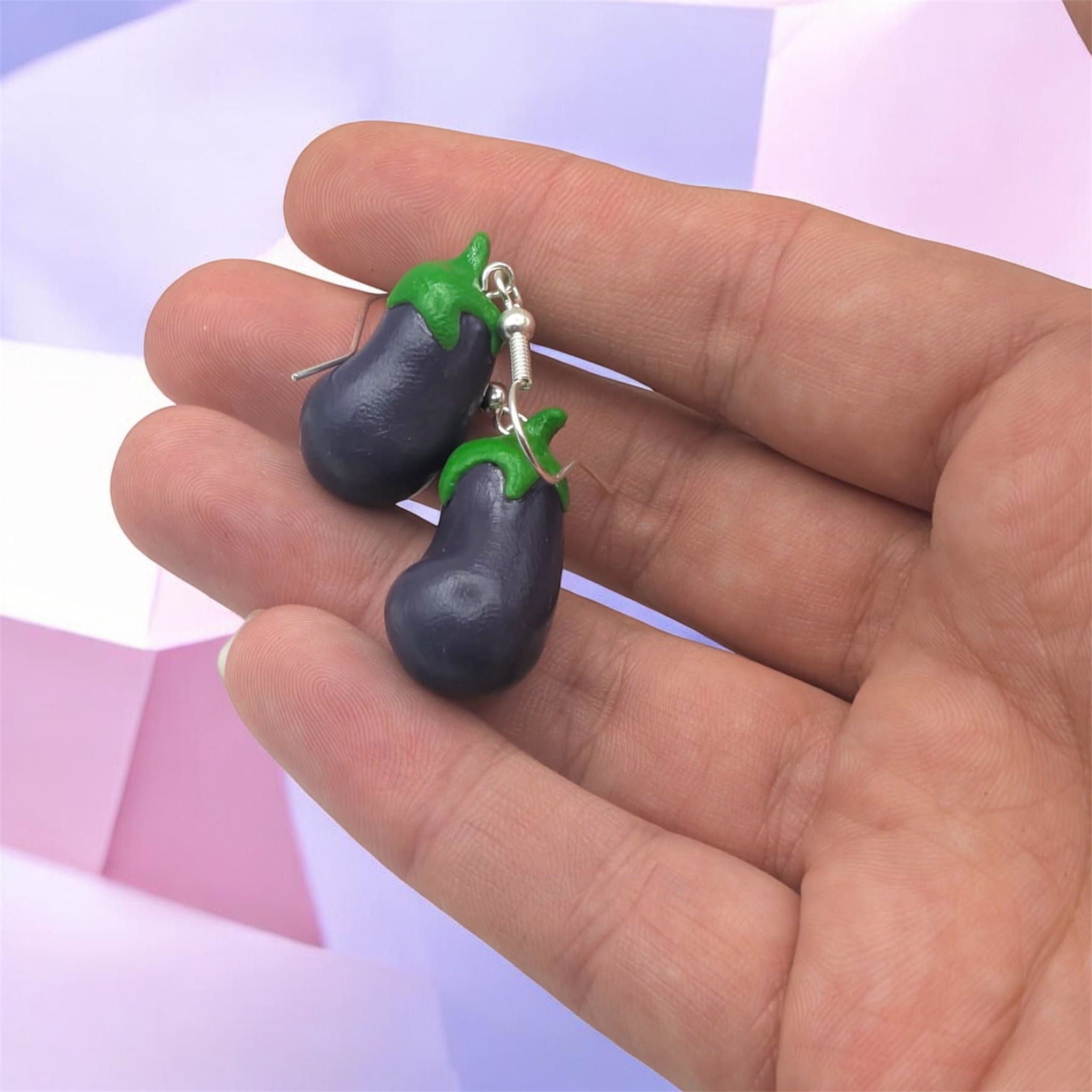 Eggplant-shaped earrings with green leaves held in a hand against a light purple background