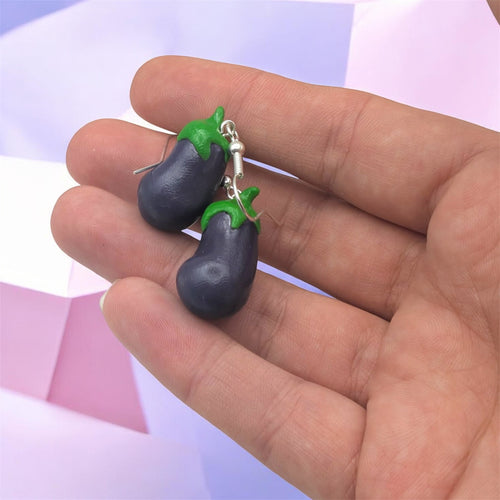 Eggplant-shaped earrings with green leaves held in a hand against a light purple background
