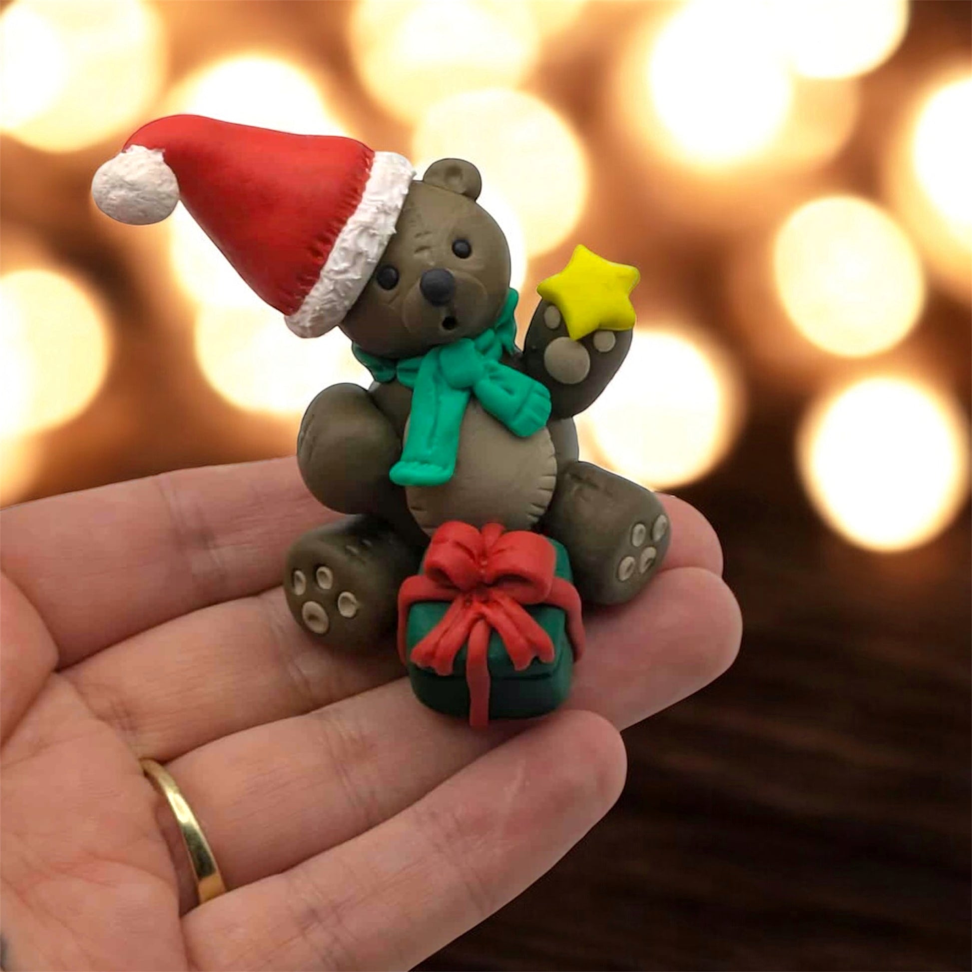 Hand holding a small bear figurine with a Santa hat, green scarf, and red bow against a blurred light background.
