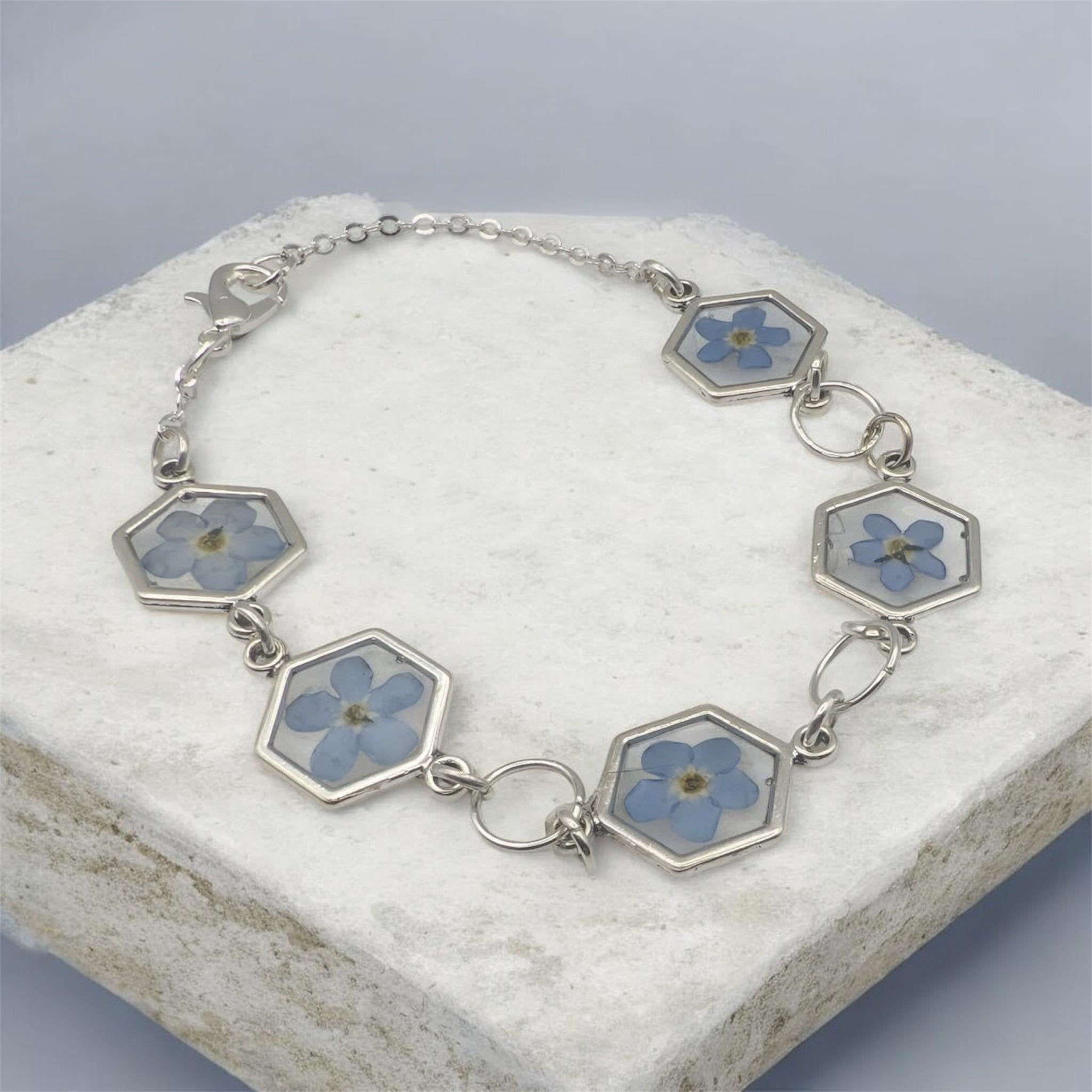 Bracelet laying flat on white surface with blue background, handmade resi. Jewellery with real pressed forget me not flowers 