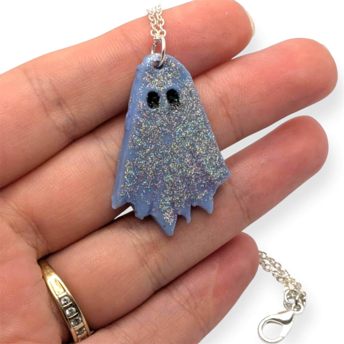 Blue ghost-shaped pendant on a chain held in a hand against a white background