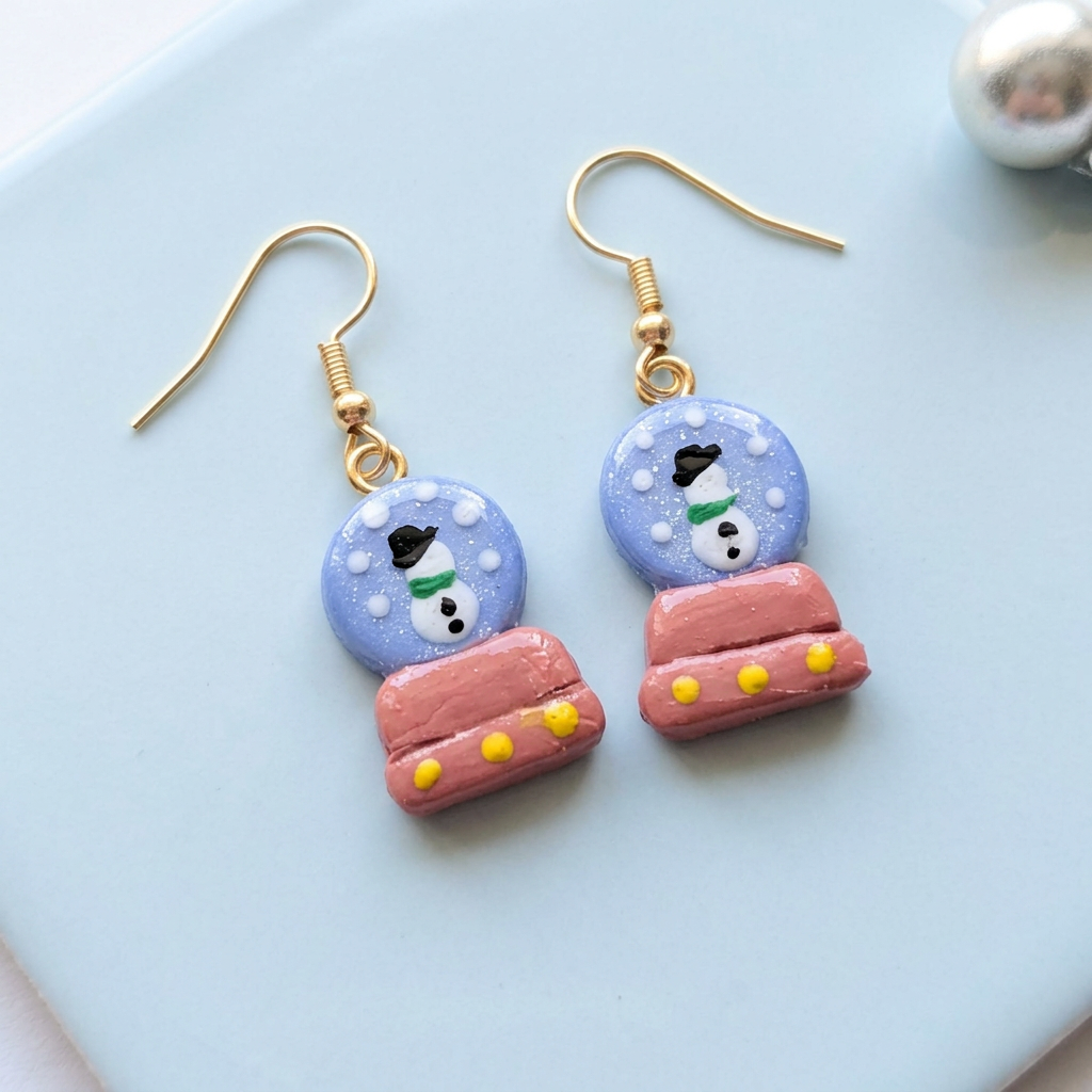 Snow globe earrings A pair of handmade dangle earrings shaped like snow globes, featuring a light blue top half with white snowflakes and a snowman face, and a pink bottom half resembling a blanket with yellow dots. The earrings have gold-tone fishhook wires and are displayed on a light blue surface.