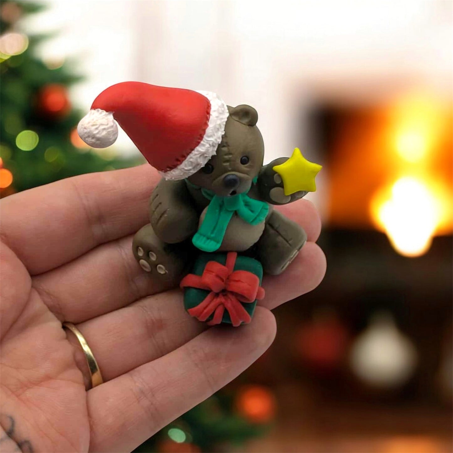 Small teddy bear figurine with a Santa hat, green scarf, and red bow held in a hand against a festive background.