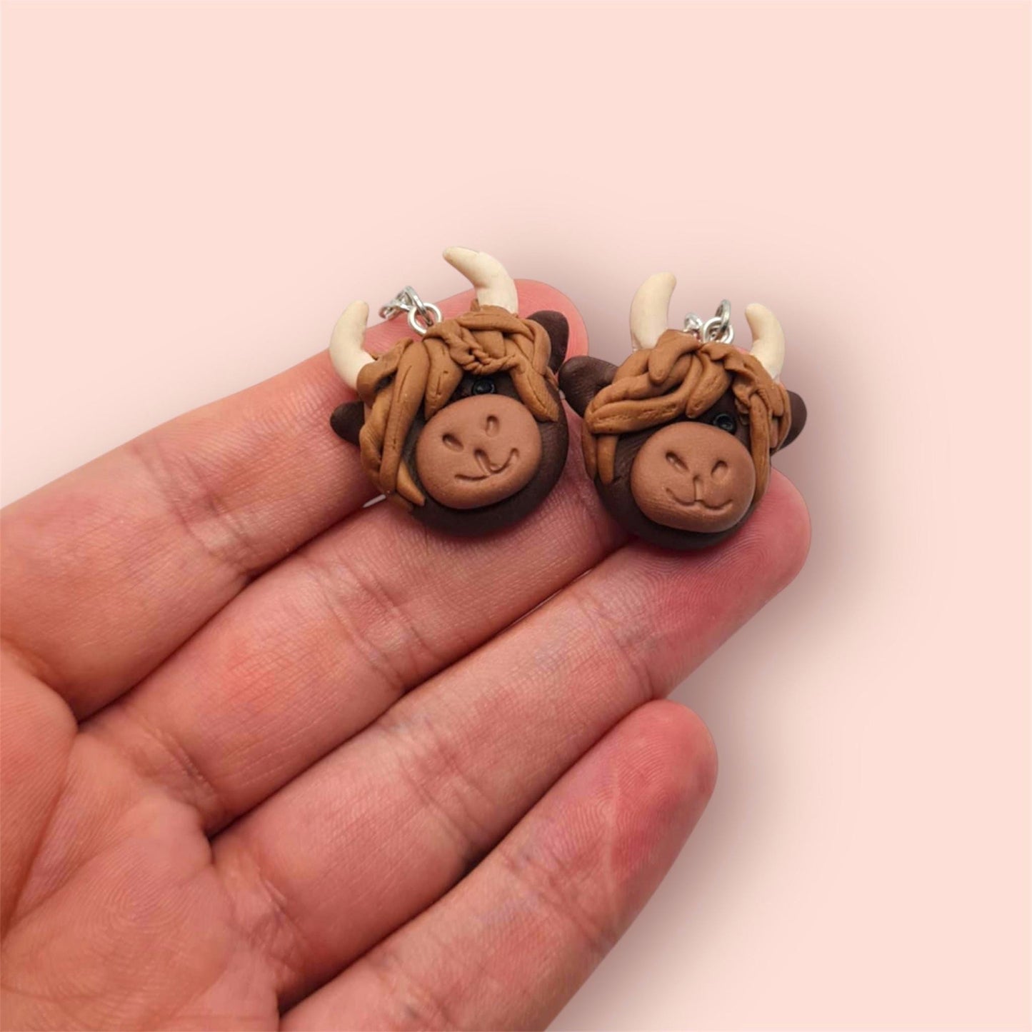 Pair of highland cow-shaped earrings held between fingers on a light pink background