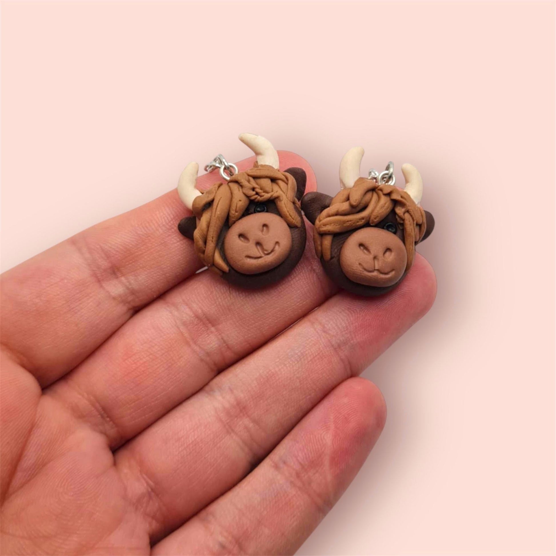 Pair of highland cow-shaped earrings held between fingers on a light pink background