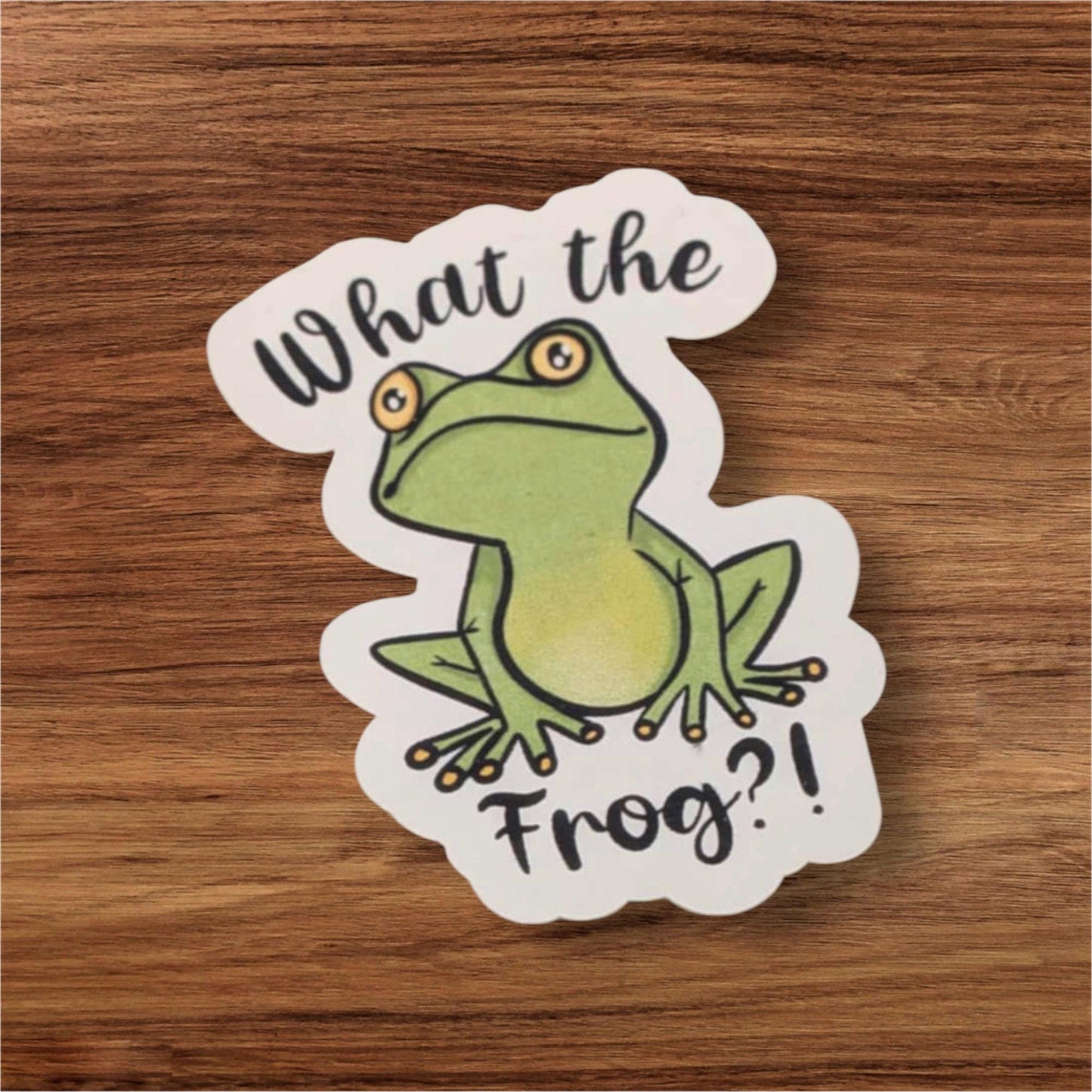 Sticker with a frog illustration and text 'What the Frog?' on a wooden surface