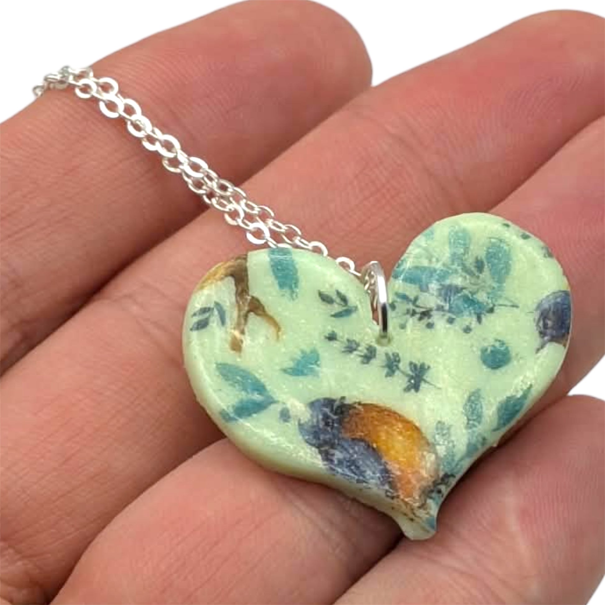 Blue bird heart necklace shown on hand on a white background 