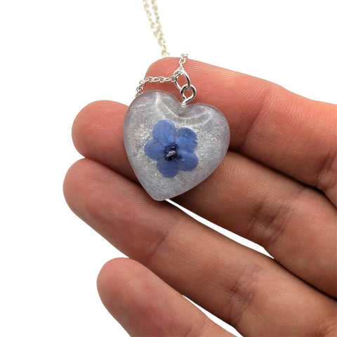 Heart-shaped pendant with a blue flower held between fingers on a white background