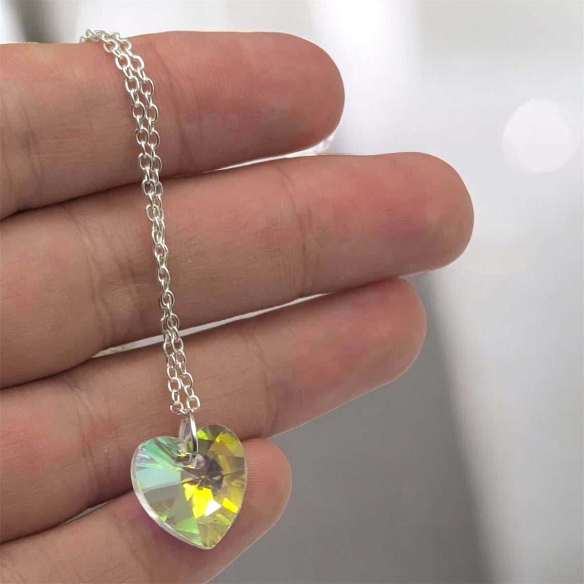 Heart-shaped crystal pendant on a silver chain held between fingers against a neutral background