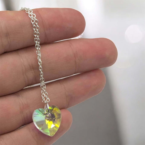 Heart-shaped crystal pendant on a silver chain held between fingers against a neutral background