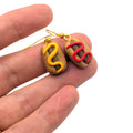Hand holding mini hot dog earrings with ketchup and mustard detail, finished with gold plated ear wires 