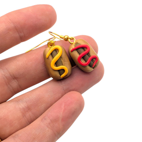 Hand holding mini hot dog earrings with ketchup and mustard detail, finished with gold plated ear wires 