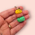 Hand holding a small, colorful charm resembling a character with a yellow face and brown and green body on a light pink background.