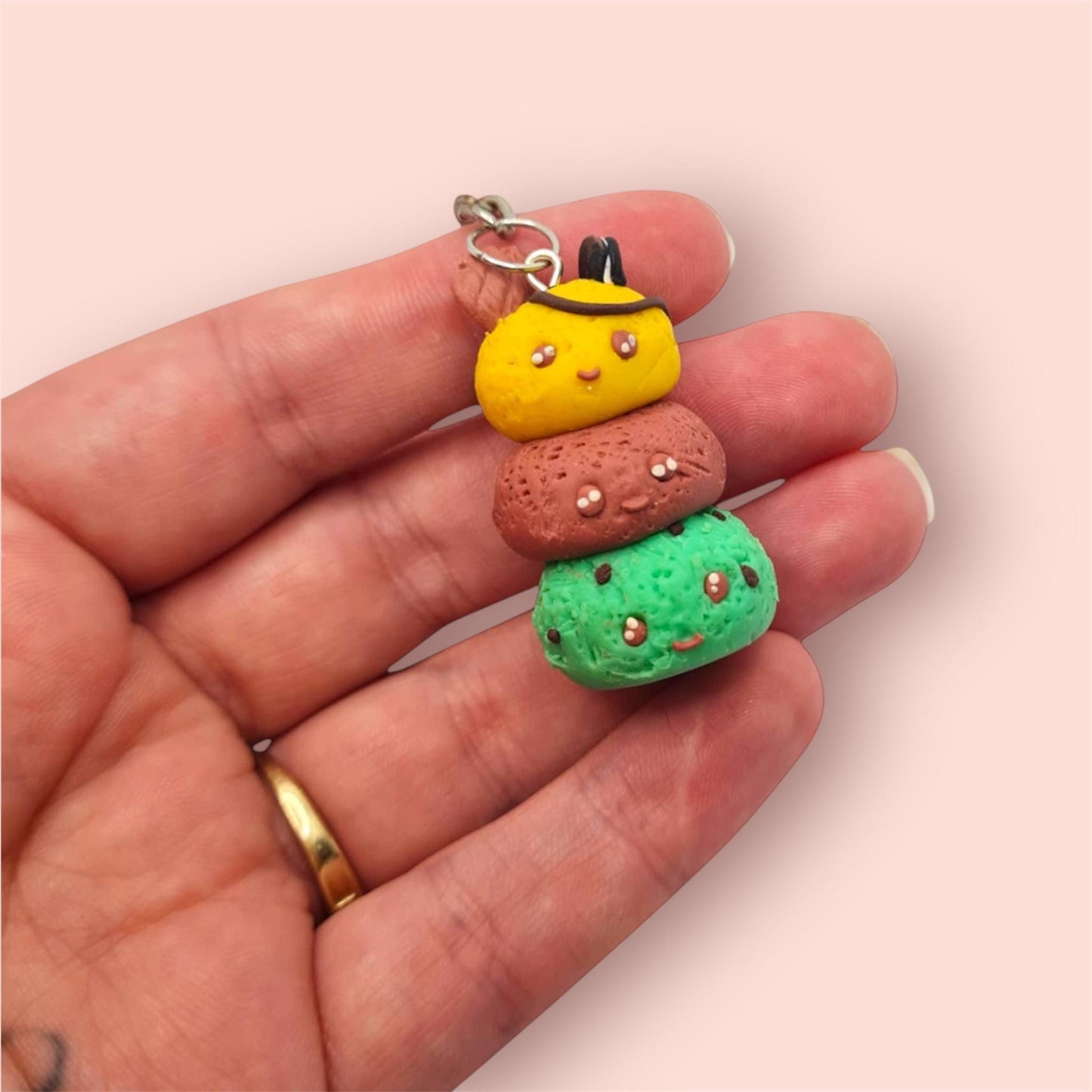 Hand holding a small, colorful charm resembling a character with a yellow face and brown and green body on a light pink background.