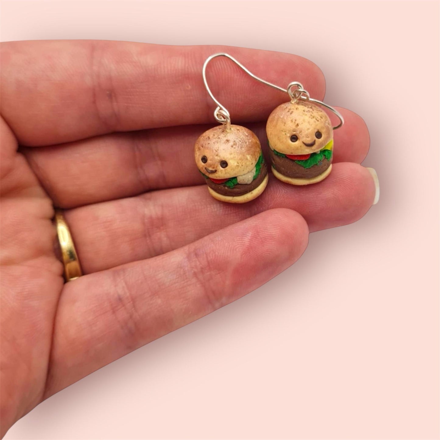 Burger-shaped earrings held in a hand on a light pink background