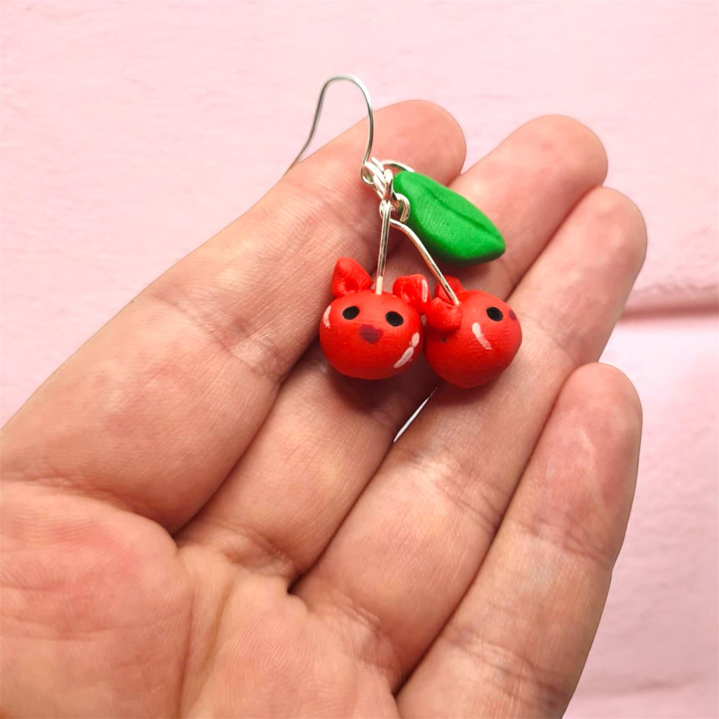 Red cherry earrings with a green leaf held in a hand against a pink background