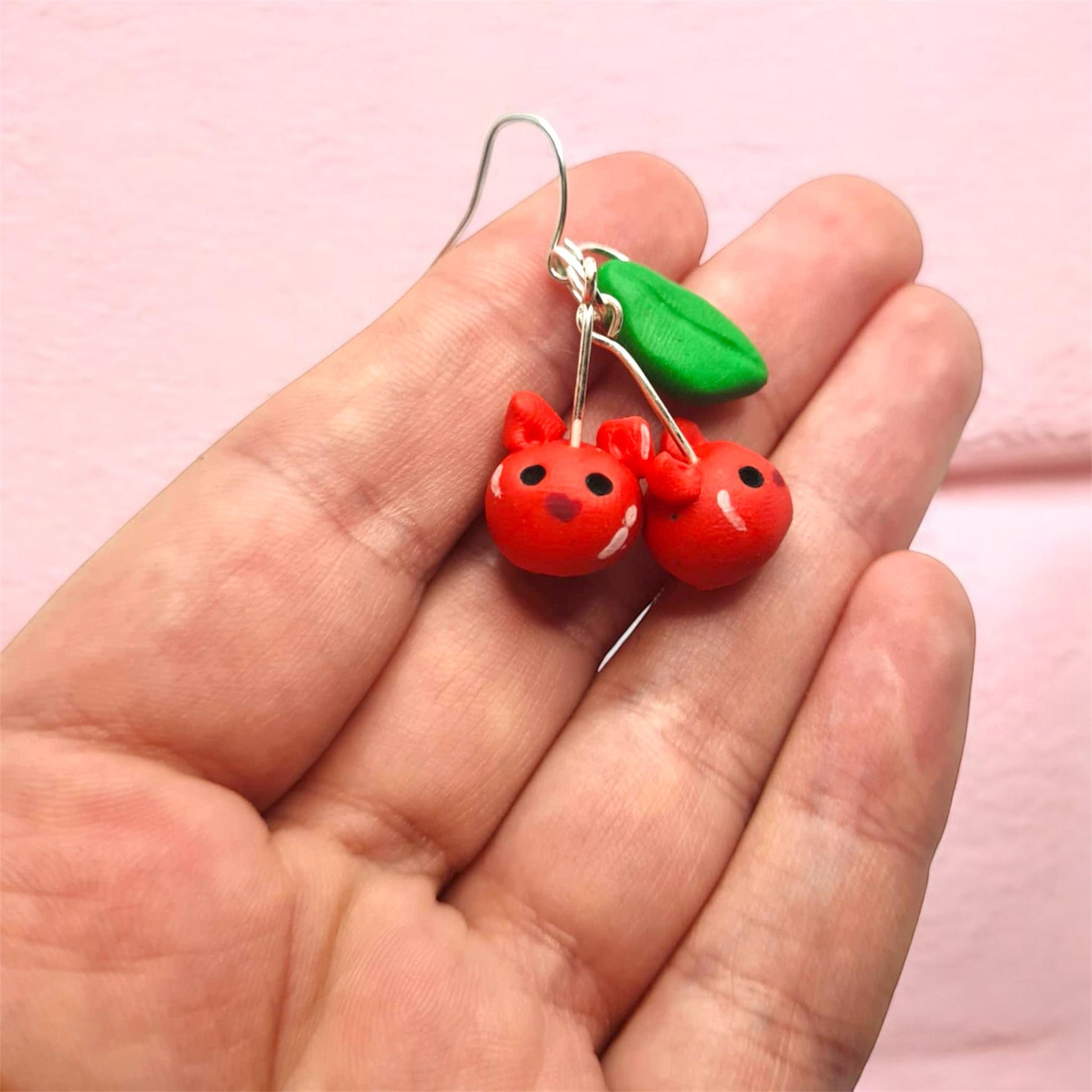 Red cherry earrings with a green leaf held in a hand against a pink background