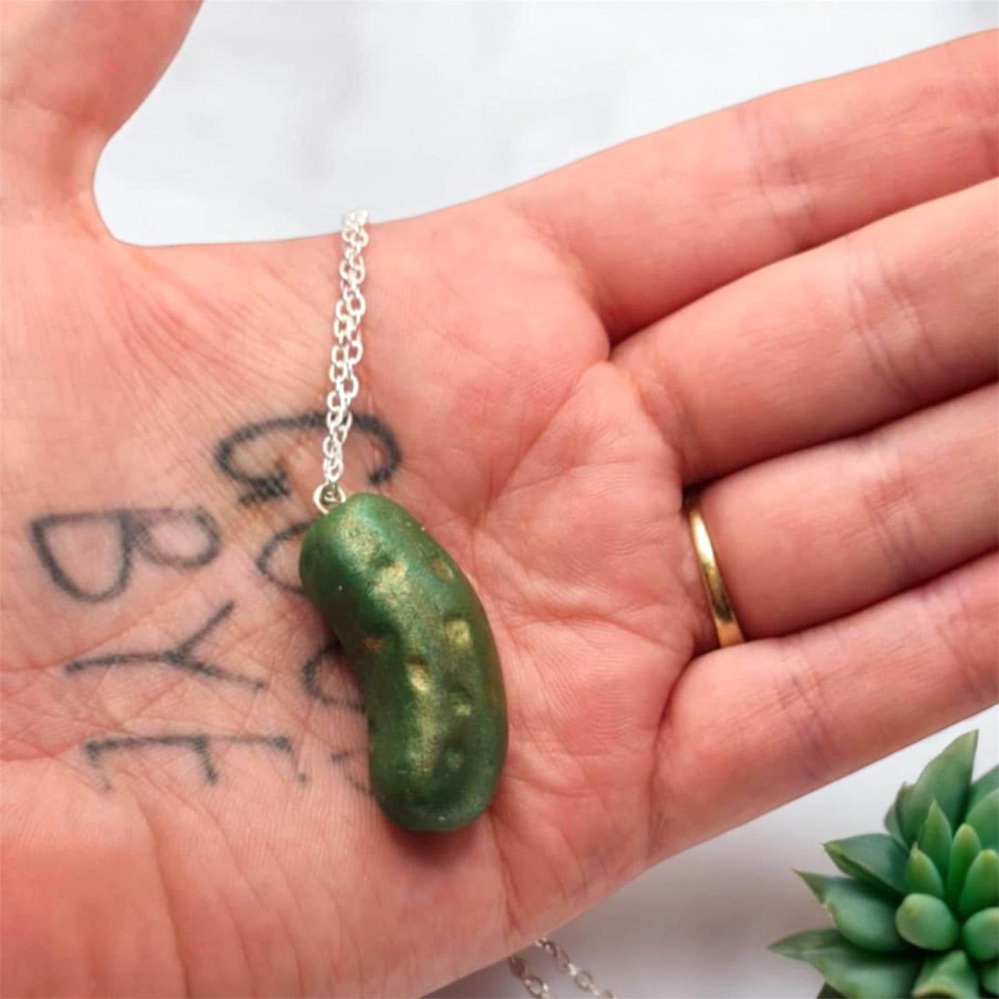 Green bean-shaped pendant on a chain held in a hand with tattoos and a ring.
