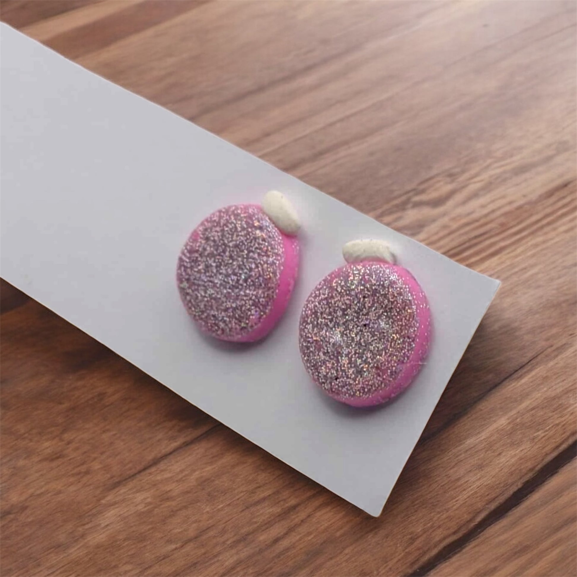 Pink earrings with glittery finish on a white card, placed on a wooden surface.