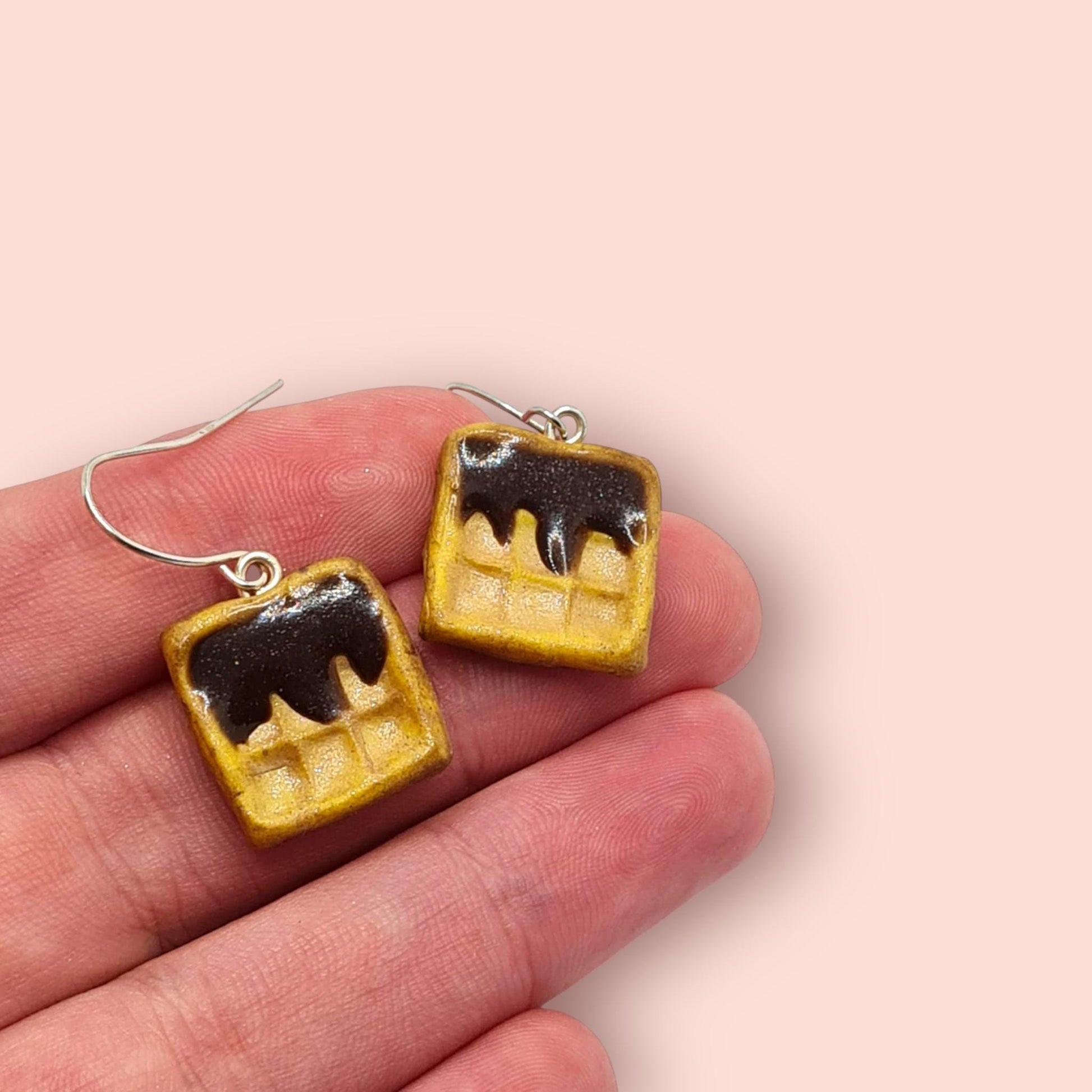 Waffle-shaped earrings with chocolate sauce on a hand against a light pink background