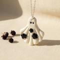 Ghost-shaped pendant necklace on a beige background with dried flowers.
