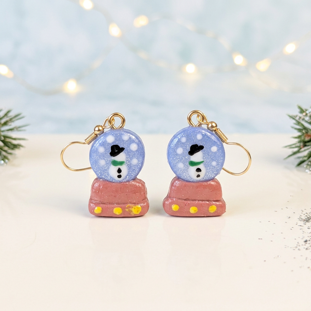 Pair of blue snow globe earrings with snowmen and pink bases on an ivory surface
