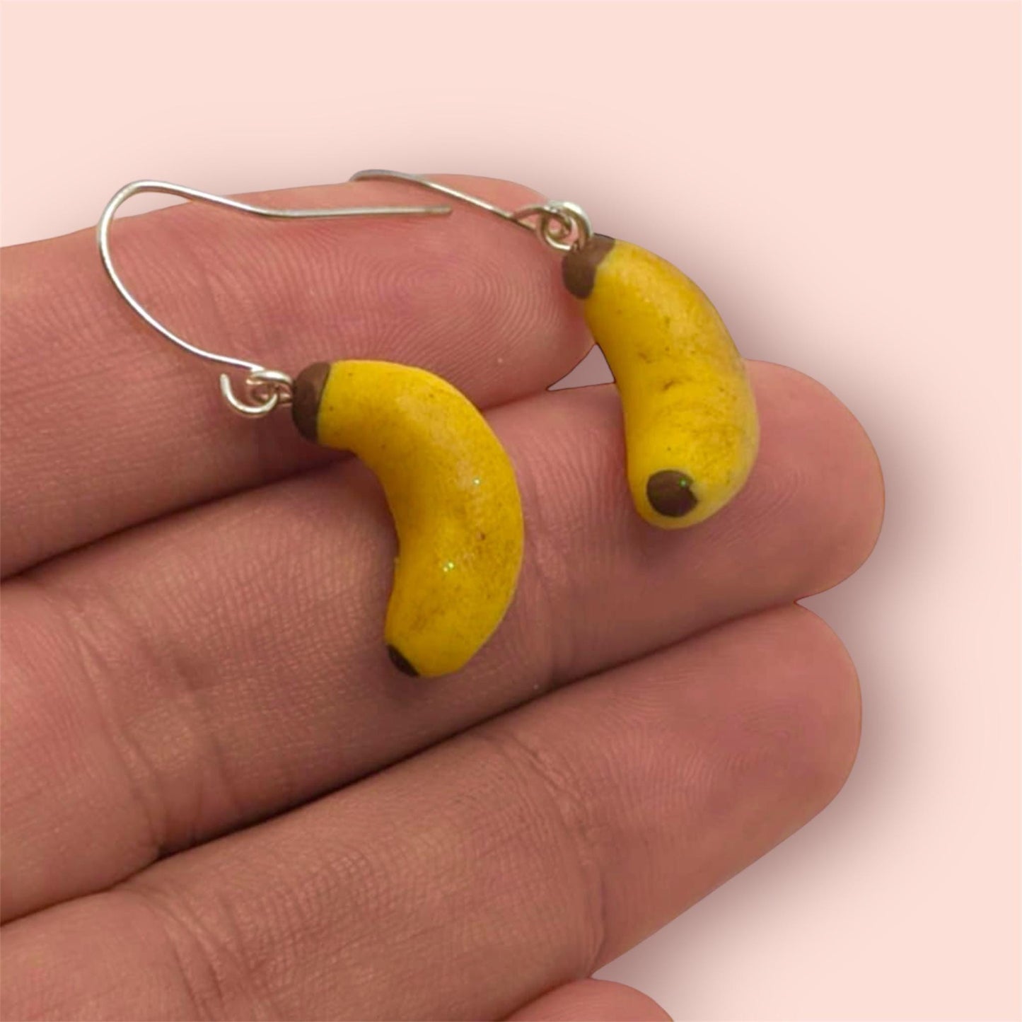 Banana-shaped earrings held between fingers on a light pink background